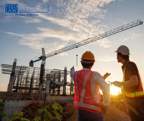 Two construction workers in hard hats and high-visibility vests stand on a dirt site at sunset, looking toward a crane and rebar framework for a building under a partly cloudy sky.