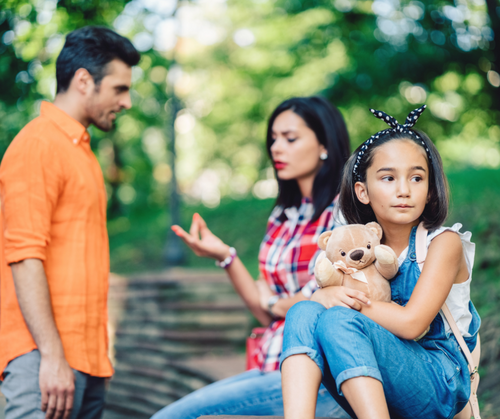 In a green park, a child in a blue dress sits on a bench holding a teddy bear with a tiara headband while an adult in an orange shirt stands nearby and a woman in a plaid shirt looks on.